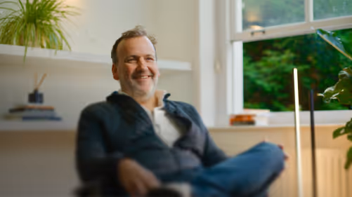 Man zit ontspannen en glimlacht in een lichte kamer met een raam en groene planten op de achtergrond.