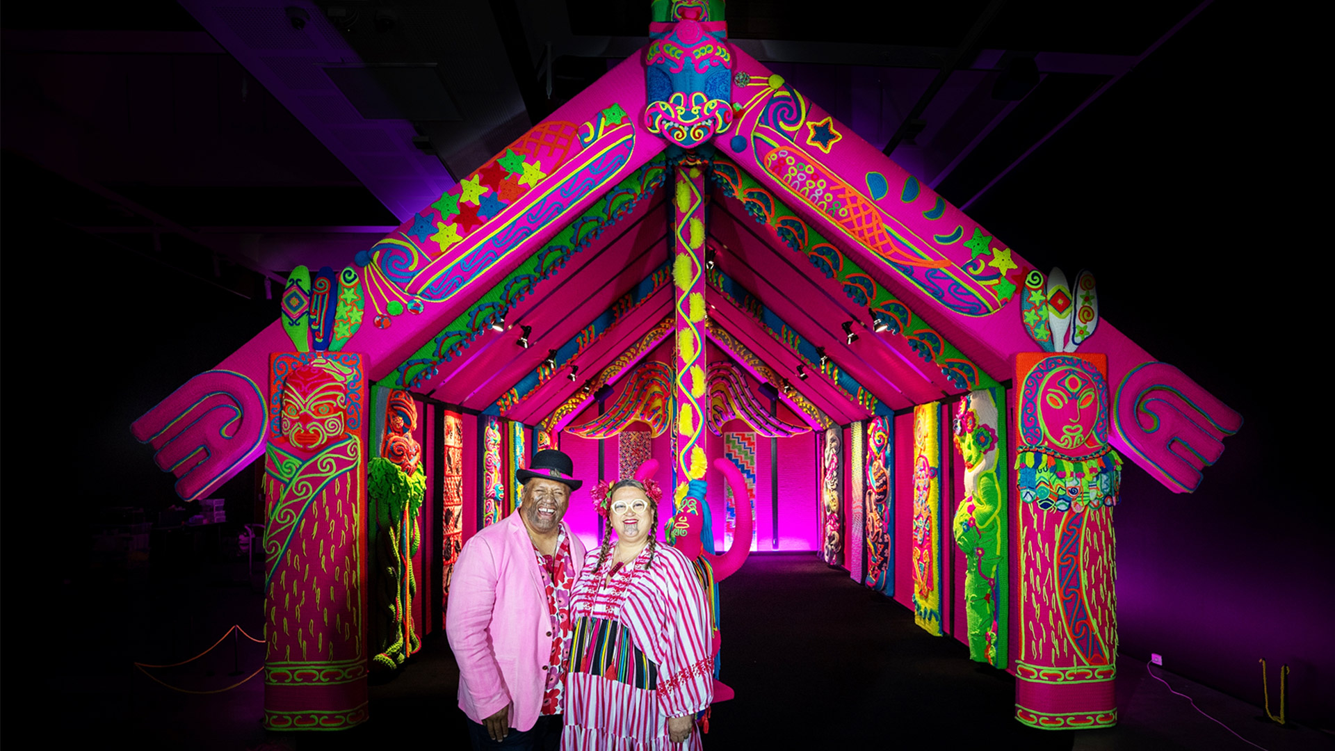 A Marae meeting house, A house of Joy, crocheted and knitted by Lissy and Rudi as featured in The Civic, The Spinoff, and Meta. Artists Lissy and Rudi Robinson-Cole stand proudly in front of Wharenui Harikoa – their radiant crocheted meeting house that transforms ancestral forms into a kaleidoscope of neon colour. Dressed in vibrant hues that mirror their creation, the pair embody the kaupapa of joy, pride and aroha that pulses through every stitch. This image captures not just the artwork, but the living spirit of its makers – a bold celebration of whakapapa, whanaungatanga, and the boundless possibilities of Māori creativity.
