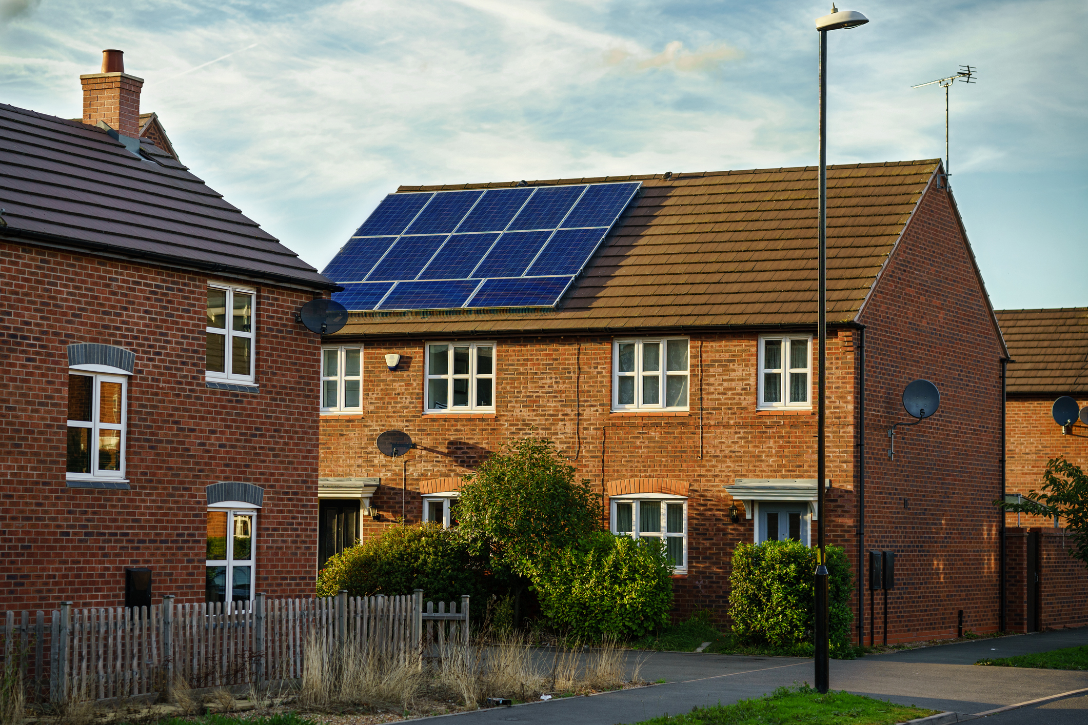 Two attached brick houses with white-framed windows, one with solar panels on the roof, surrounded by green bushes and a wooden fence under a partly cloudy sky.
