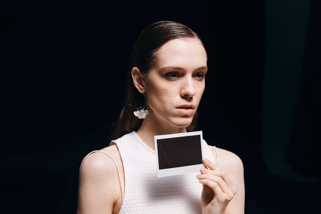 Woman in white sleeveless top holding a blank instant photo against a dark background.
