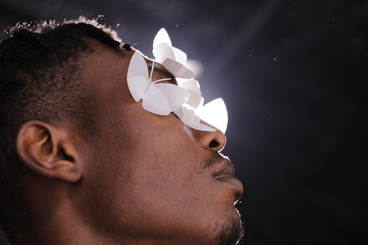 Close-up side profile of a man with white paper butterflies covering his eyes against a dark background.