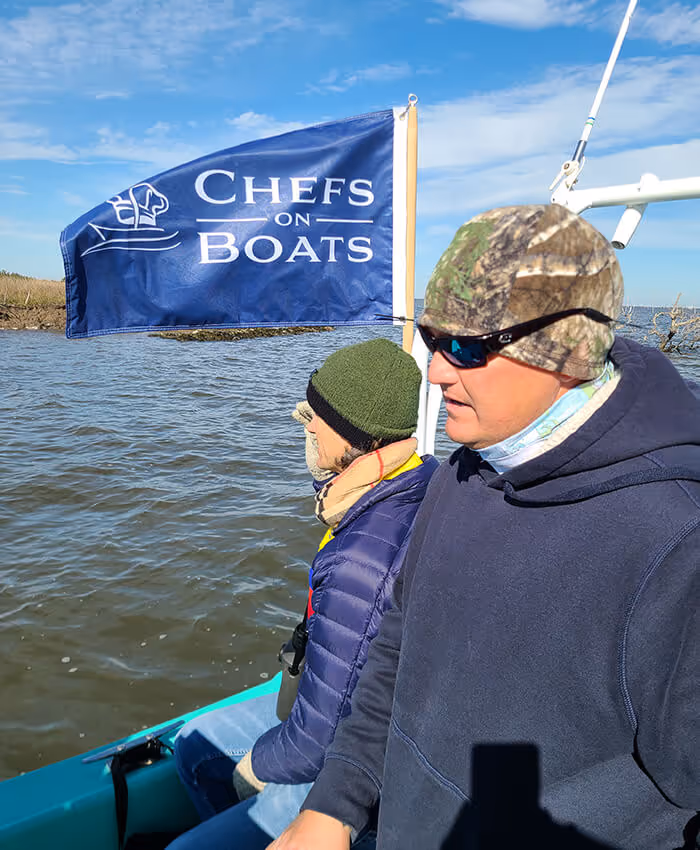 man in a hat and sunglasses on a boat with a woman in the background and a Chefs on Boats flag