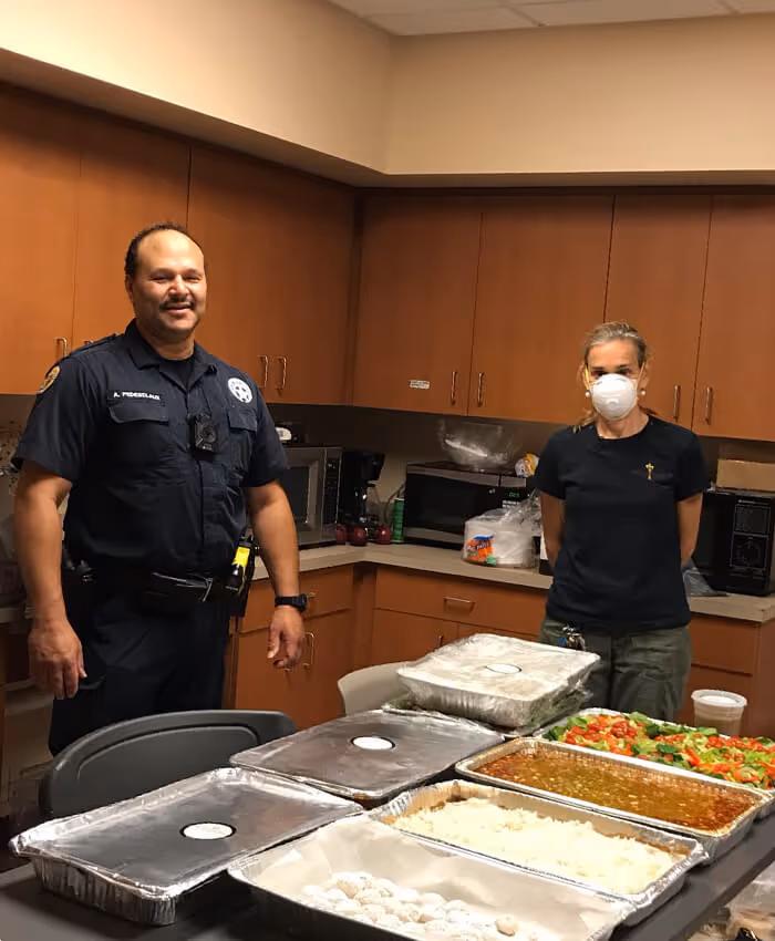 a first responder in uniform standing next to a woman in a mask by a table with containers full of food