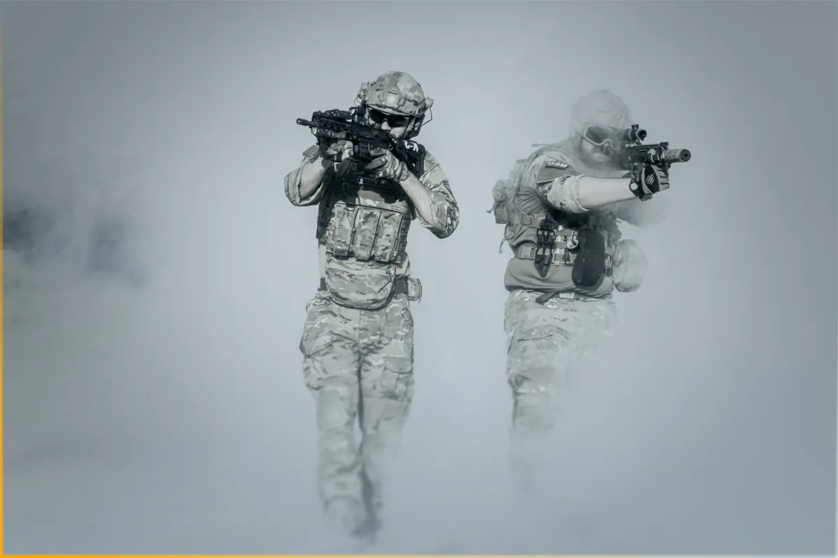Two soldiers in camouflage gear aiming rifles, moving forward through smoke or fog.