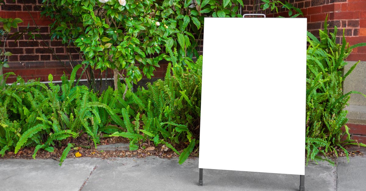  A white, blank A-frame sign is situated on a gray sidewalk. Green plants and a brick building are in the background.