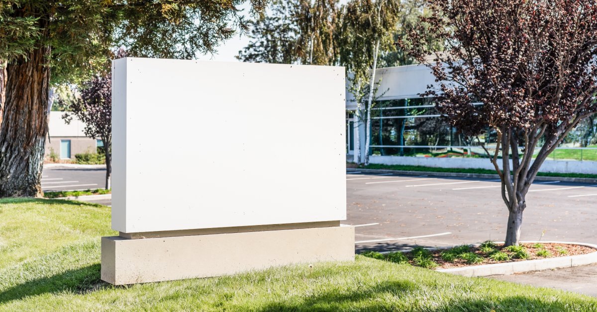 A blank, white sign is on a concrete base on a patch of grass. A parking lot and a building are in the background.