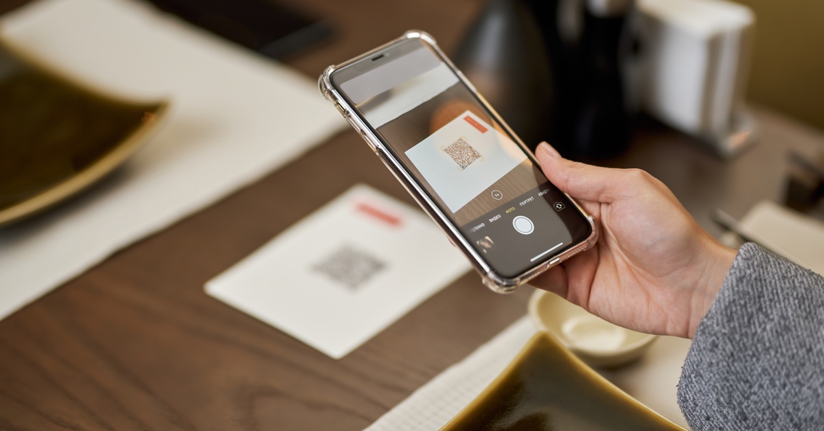 A person holds a phone with the camera application open. The screen displays a QR code on a piece of paper.