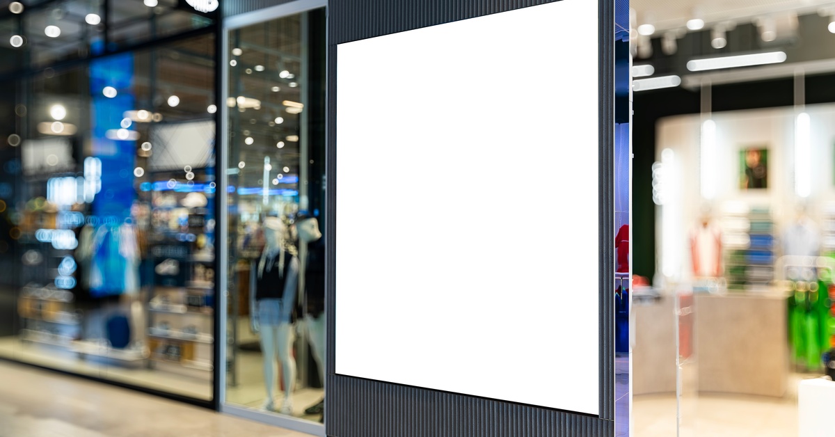 A clothing store with glass windows is blurry. On a textured gray wall, there's a blank, square, white banner.