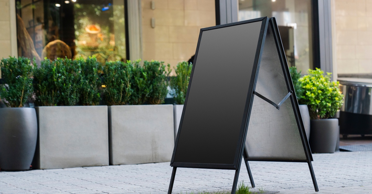 A black, blank A-frame sits on gray pavers. Storefronts with glass panels and greenery are in the background.