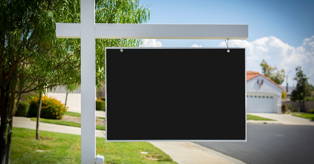 A real estate yard sign is in a neighborhood. It has a white post and a black, blank sign attached to it.