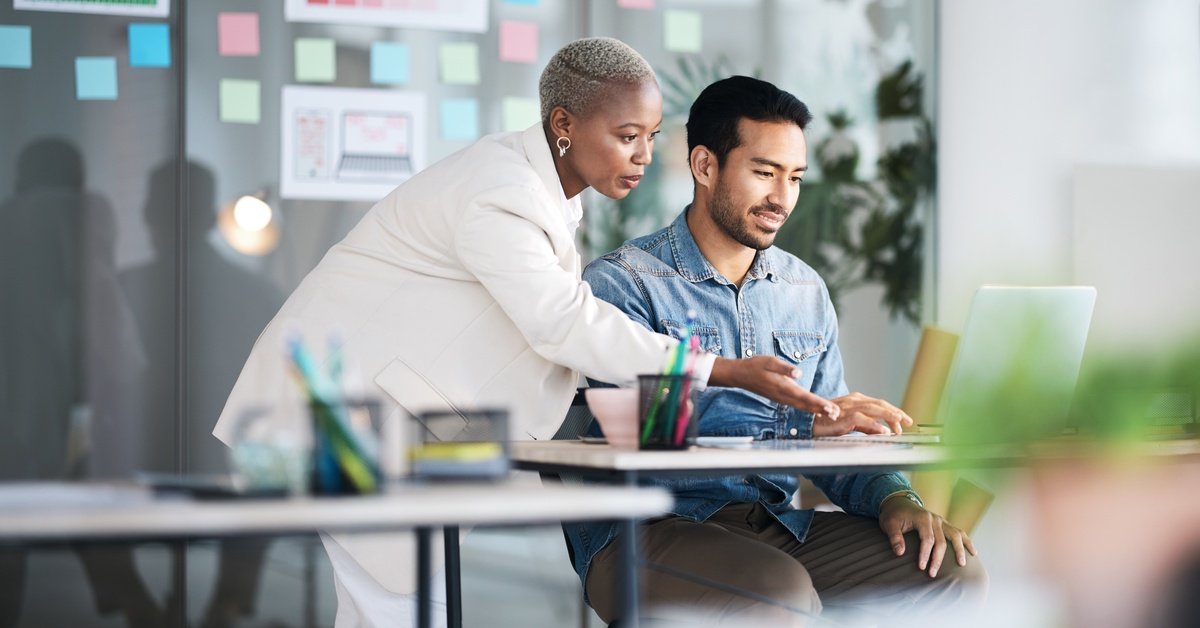 In an office, a person sits at a desk looking at a laptop as another person leans over and points at the screen.