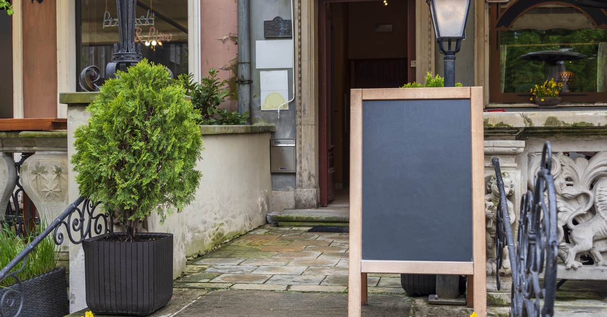 A black, blank A-frame sign is propped up in front of a storefront with a wooden door frame and concrete walls.