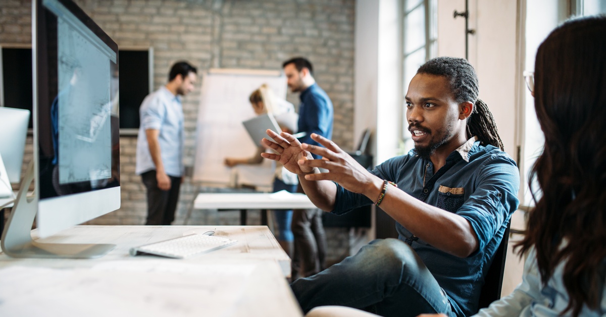 Two people sit at a desk in front of a computer monitor. A man speaks with both of his hands outstretched.
