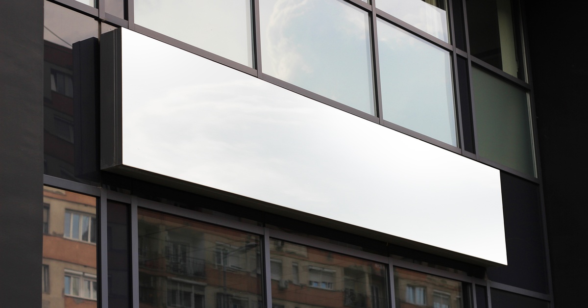 A building with square, glass windows has a rectangular sign that's white and blank. The windows reflect a brick building.