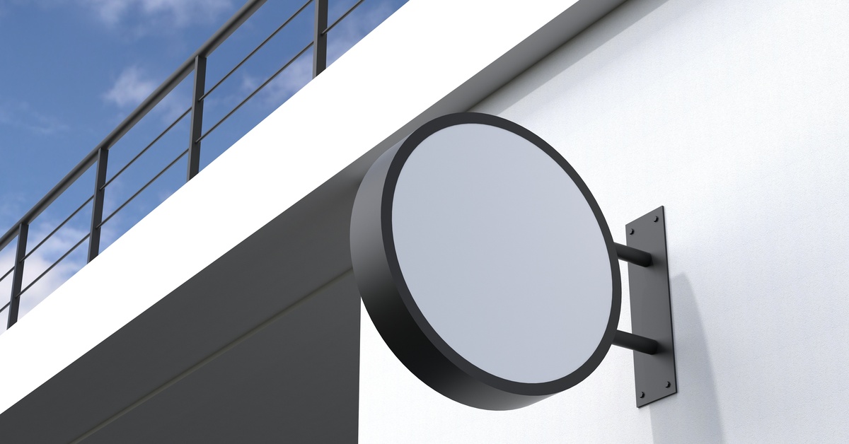 A small, round sign with black edges and a white center is mounted on a white building. The roof has a black railing.