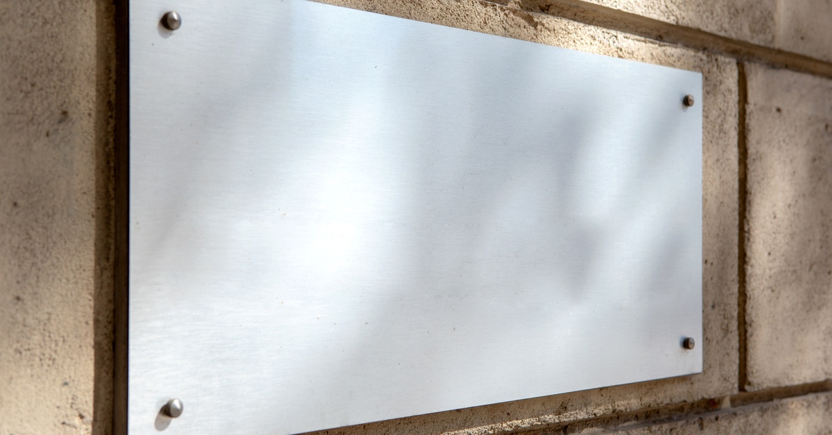 A close-up of a rectangular, blank, white sign. The sign is bolted to a tan wall made of large bricks.