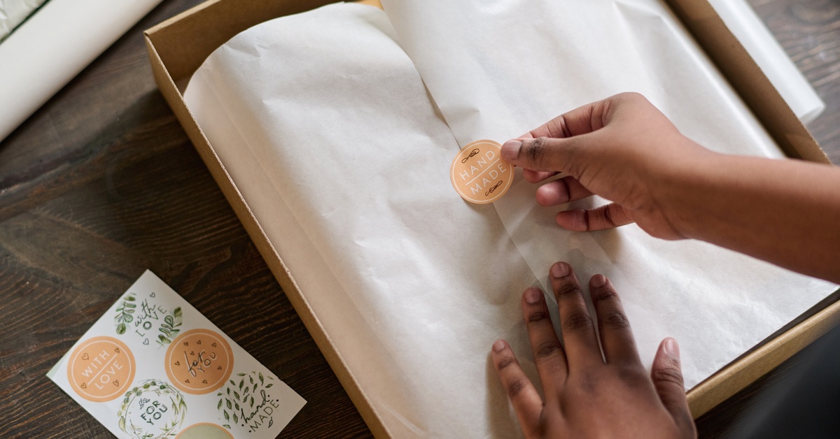 A person places an orange sticker that reads, 'HANDMADE,' on an item covered with white tissue paper.