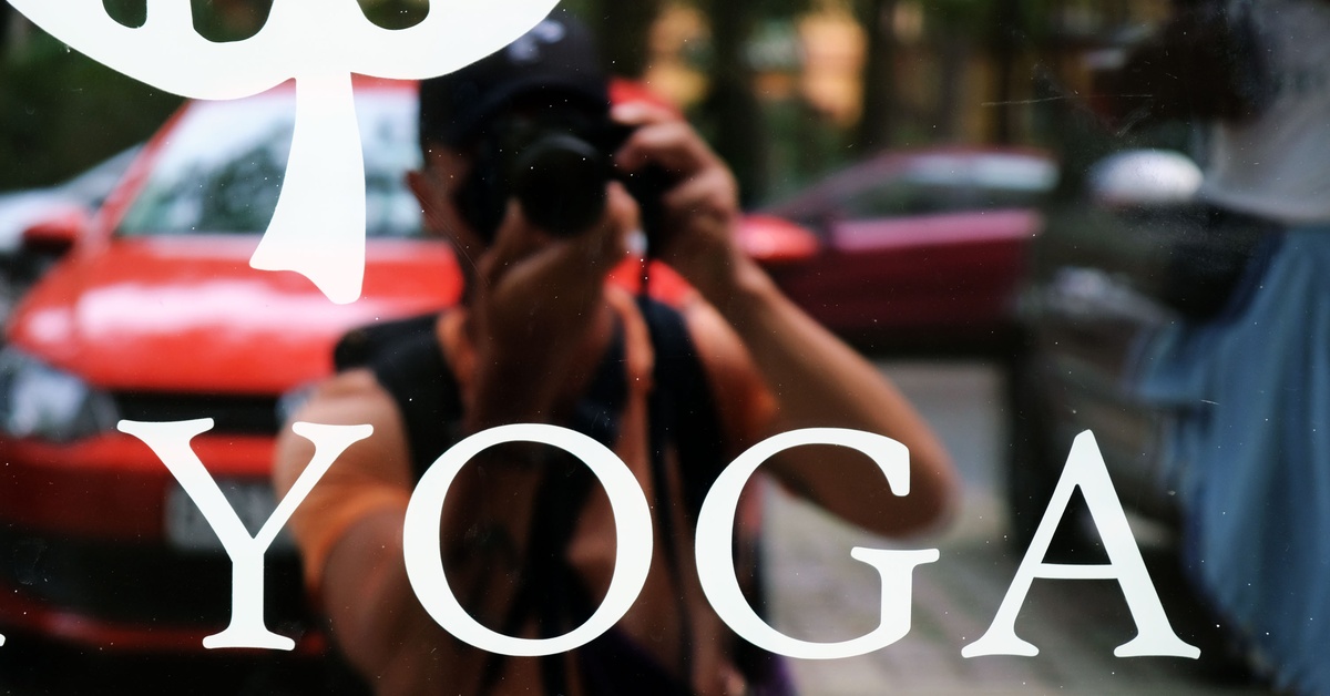 The reflection of a person in a window holding a camera and taking a photo. The window has a white decal that reads, 'YOGA.'
