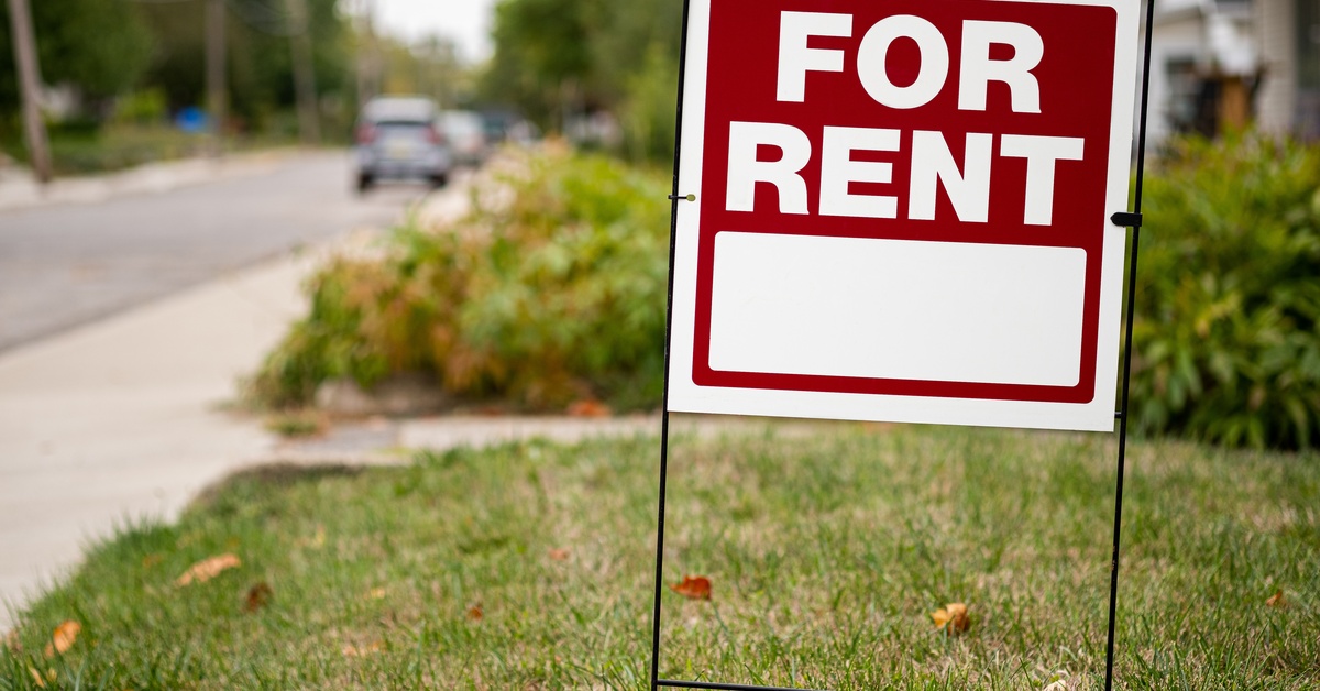 A close-up of a metal red and white "FOR RENT" sign staked in a yard. Fallen leaves are scattered in the grass.