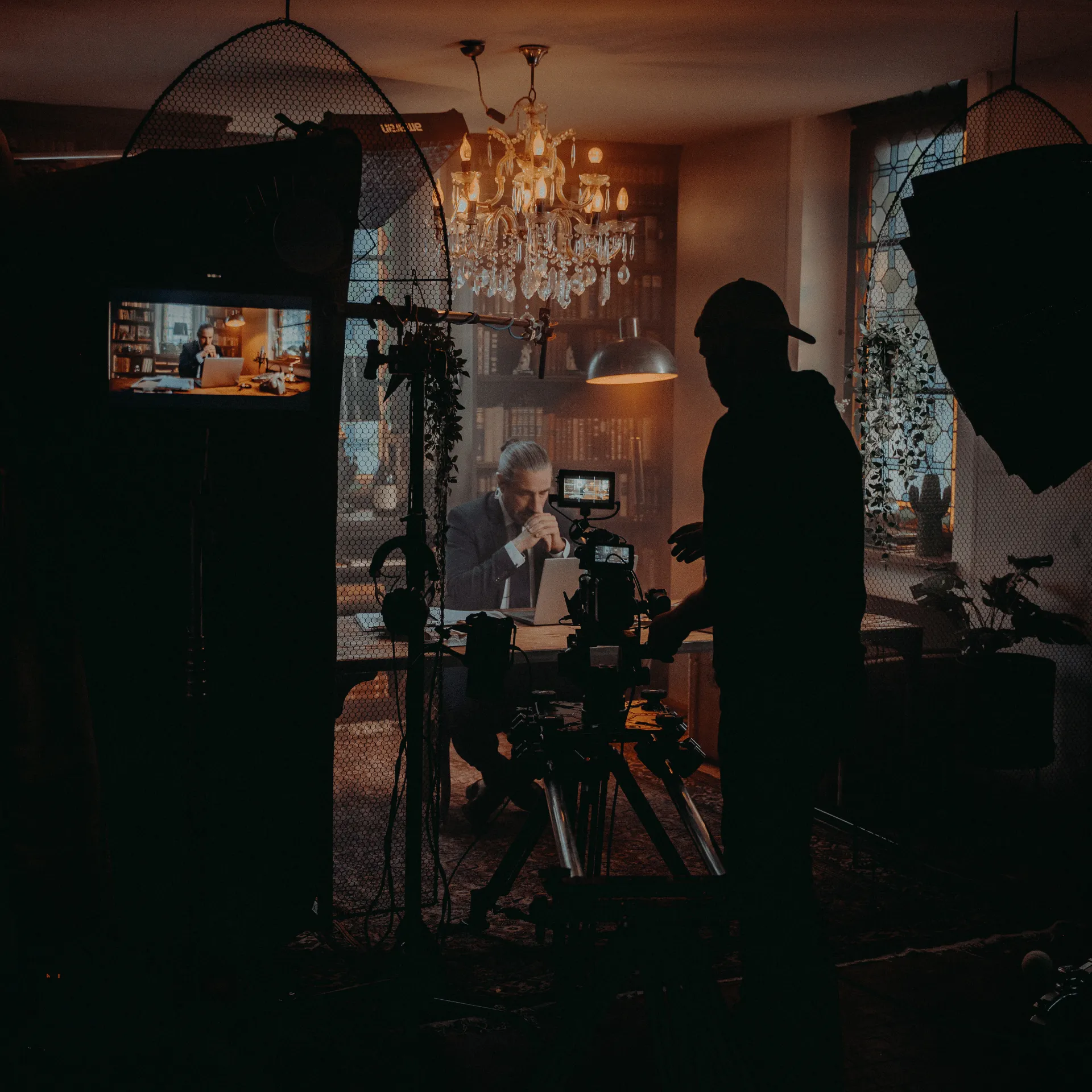 Film crew filming a man in a suit working thoughtfully on a laptop in a warmly lit, book-filled room.