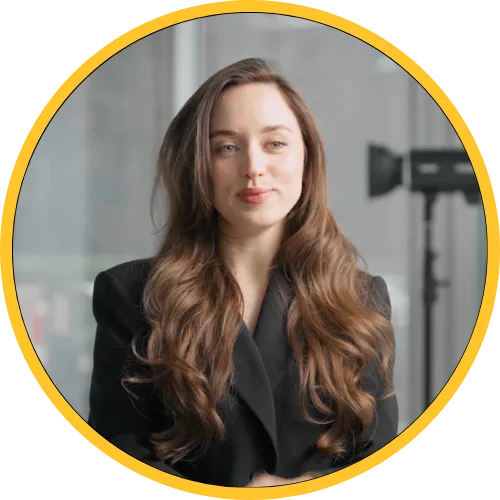 Woman with long wavy brown hair wearing a black blazer, seated indoors with blurred background.