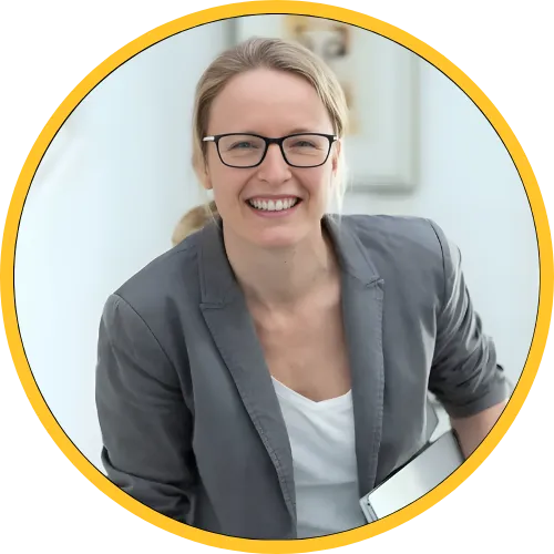 Smiling woman wearing glasses and a gray blazer, holding a laptop, in a bright indoor setting.