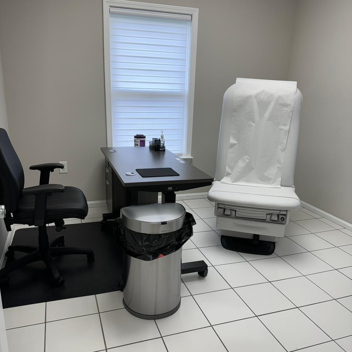 Clean medical examination room with a white exam chair covered with paper, a black office chair, a dark wood desk, and a stainless steel trash can.