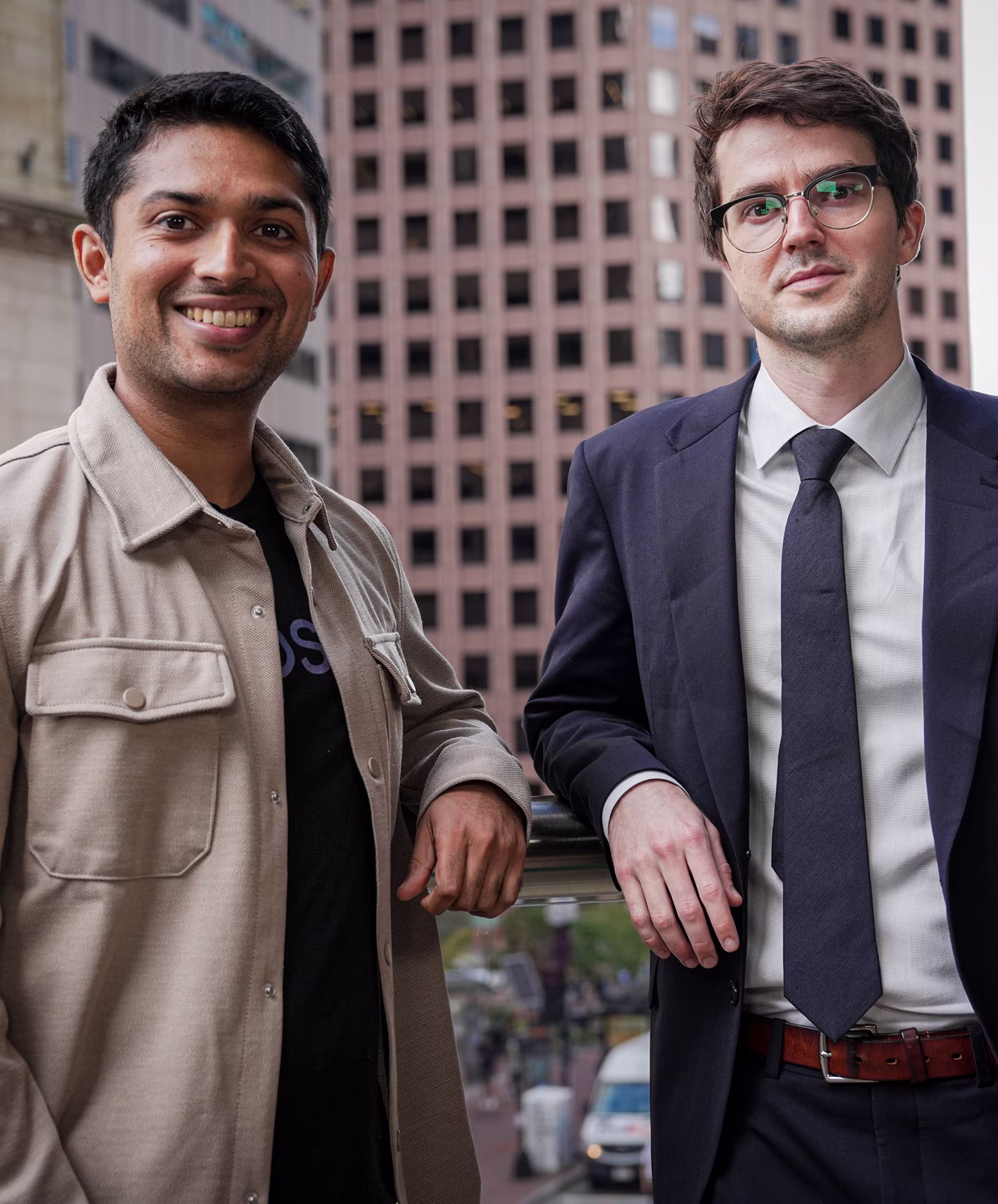Two men standing outdoors in an urban setting, one wearing a beige jacket smiling and the other in a dark suit with glasses looking at the camera.