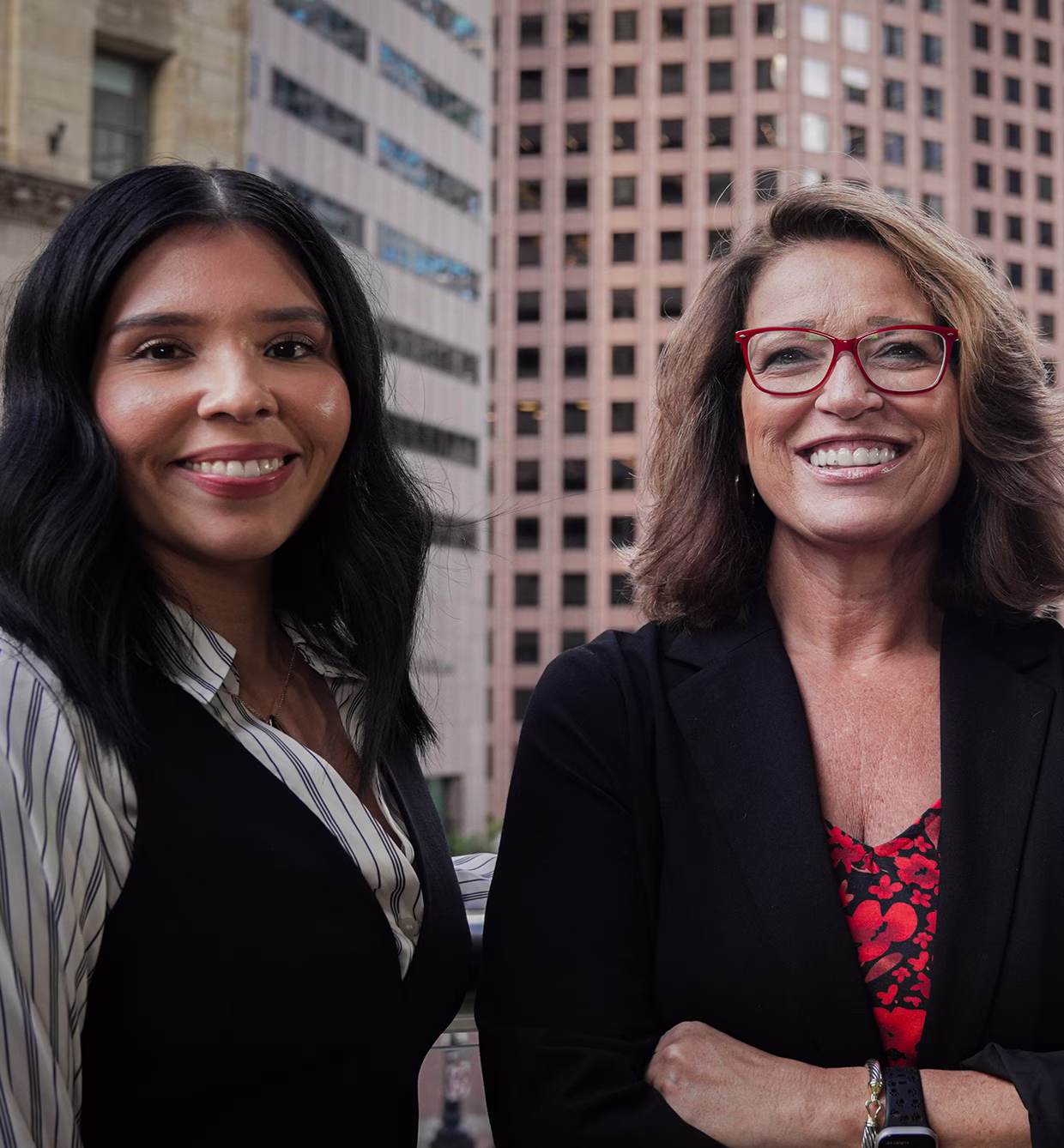 Two professional women smiling outdoors with city buildings in the background.