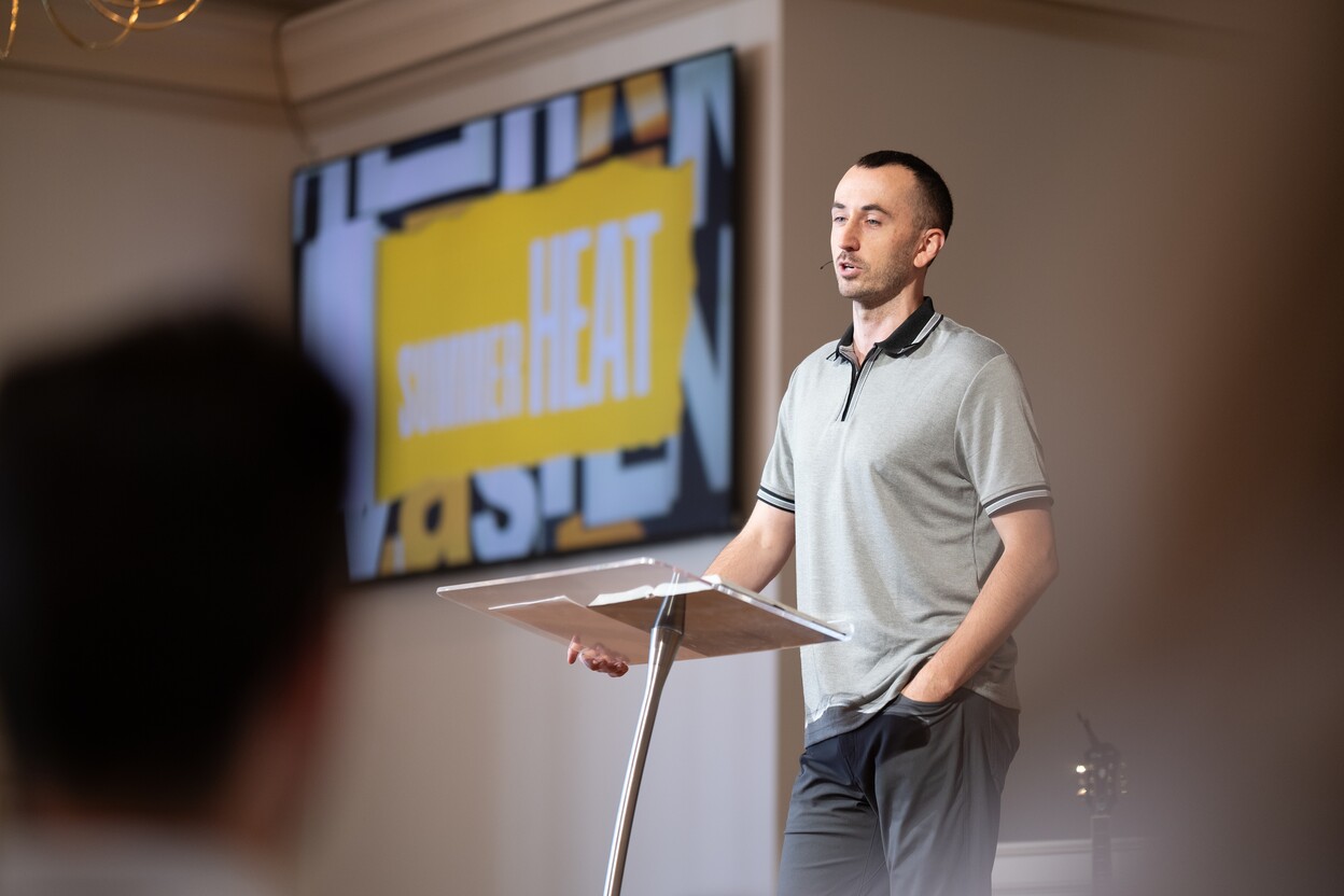 Man speaking at a podium with a screen behind him showing text 'SUMMER HEAT'.