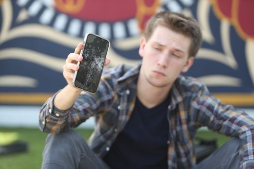 A young man holding up a phone with a shattered screen looking sad