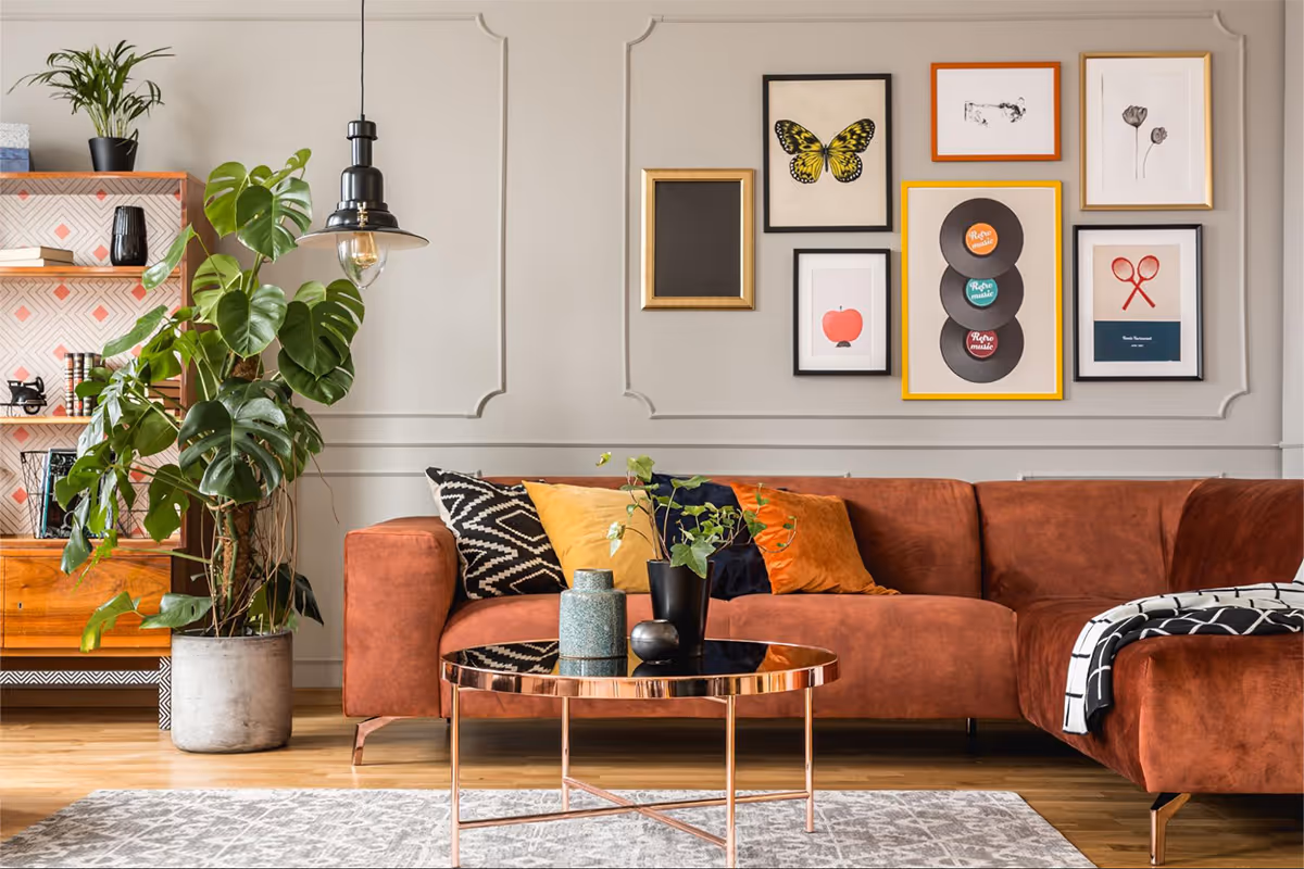 Modern living room with a rust-colored sofa, colorful cushions, a copper coffee table with plants, and a wall with framed art including a butterfly and vinyl records.