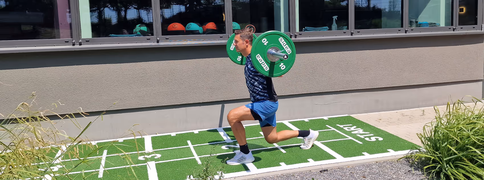 Ole Nauert, Inhaber der Rheinblick Physio, trainiert im Außenbereich der Praxis mit einer Langhantel auf einem grünen Sprint-Teppich und macht lunges.