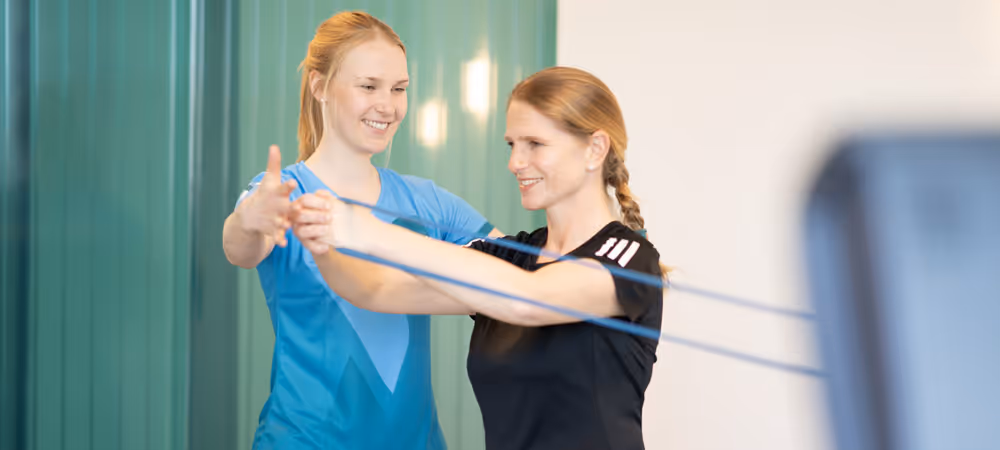 Physiotherapeutin der Rheinblick Physio trainiert mit Patientin, die eine Rückenübung mit dem Theraband durchführt.