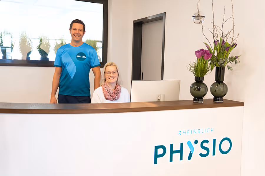 Der Inhaber und Therapeut der Rheinblick Physio steht hinter dem Empfang der Praxis. Seine Kollegin vom Empfang ist ebenfalls hinter dem Empfang und sitzt auf ihrem Stuhl.