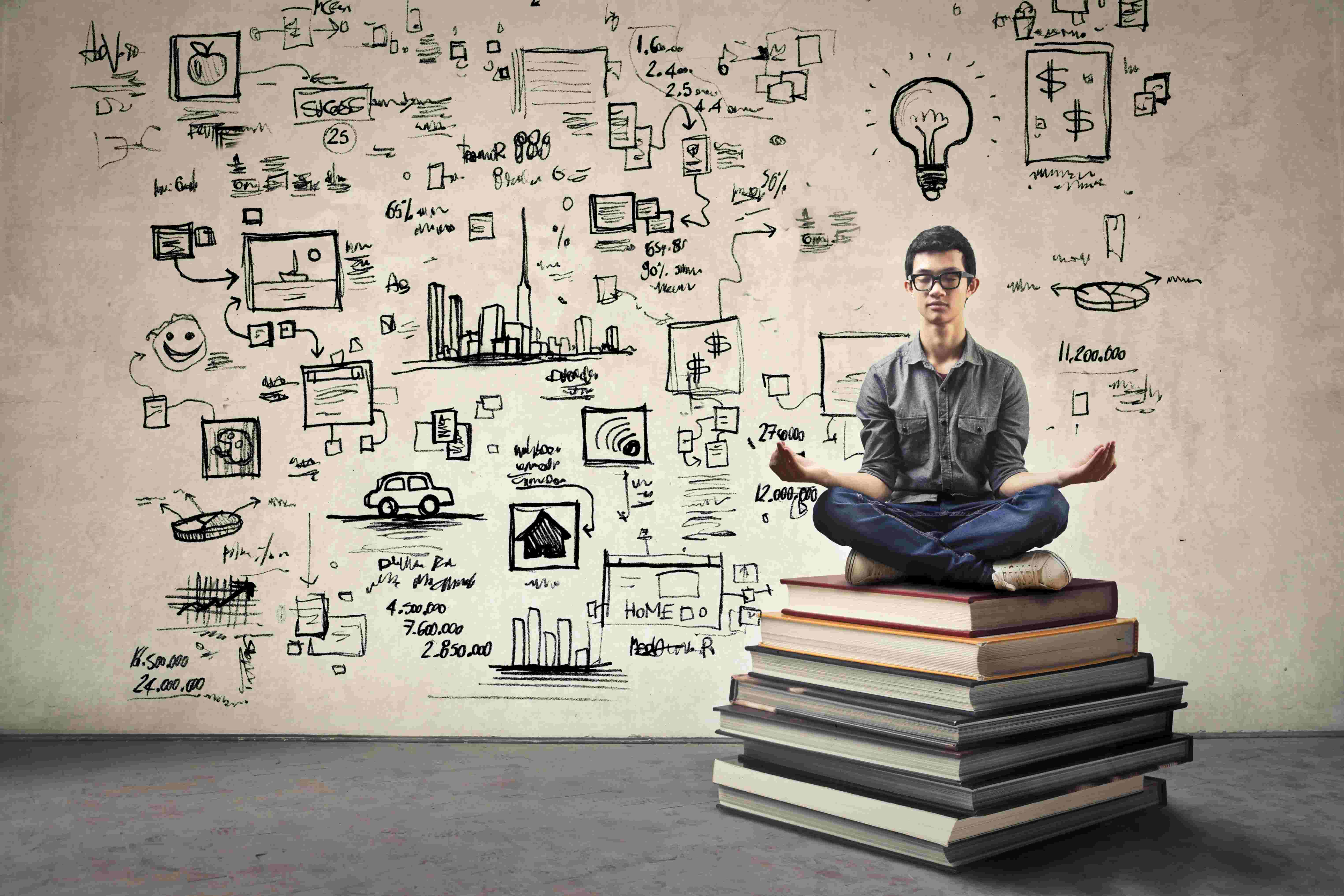 Man meditating on book stack, drawings on wall.