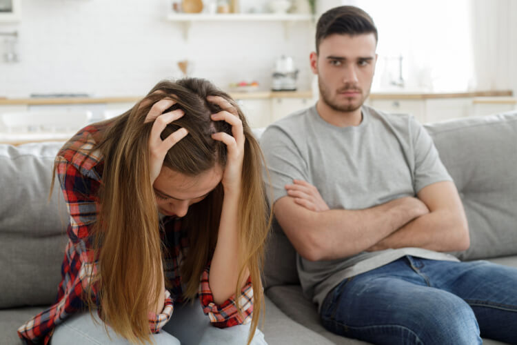 Upset woman with head in hands, man sitting beside her, arms crossed.
