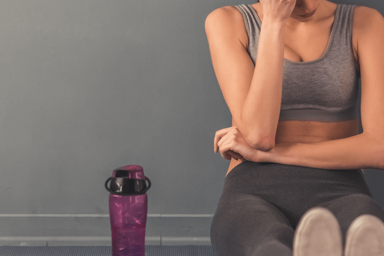 Woman sitting cross-legged in workout clothes holding her forehead, water bottle nearby.