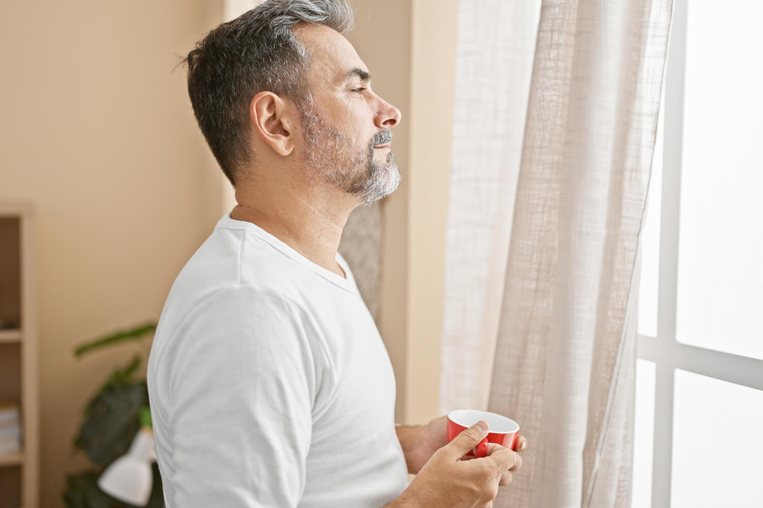 Man in white shirt holding cup, gazing out window.