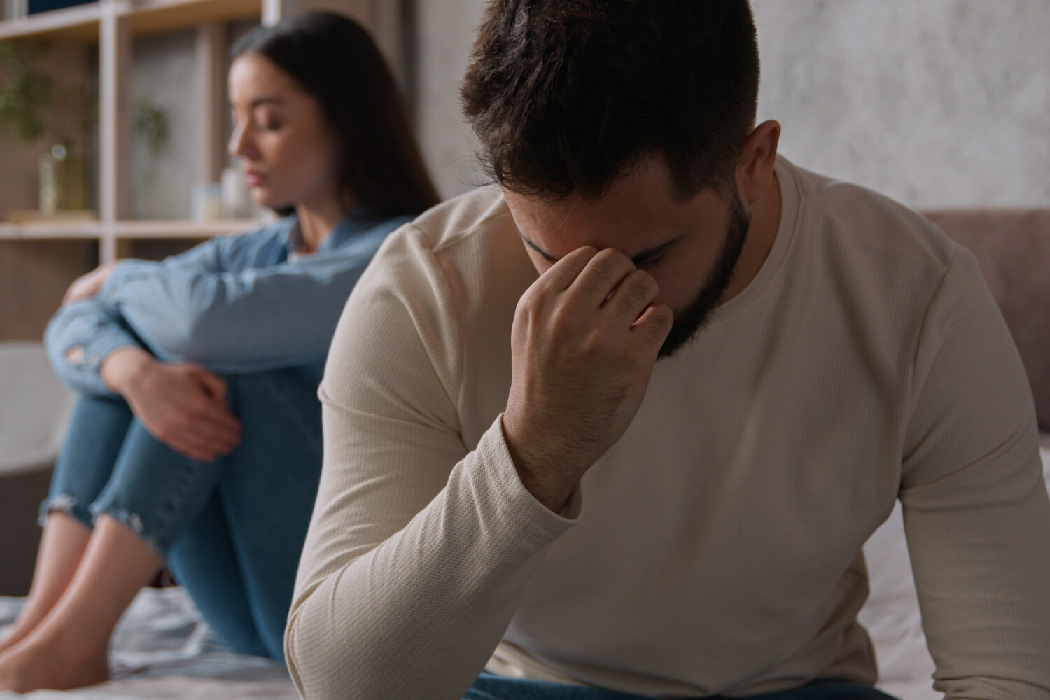 Upset man sits on bed, woman sits in background.