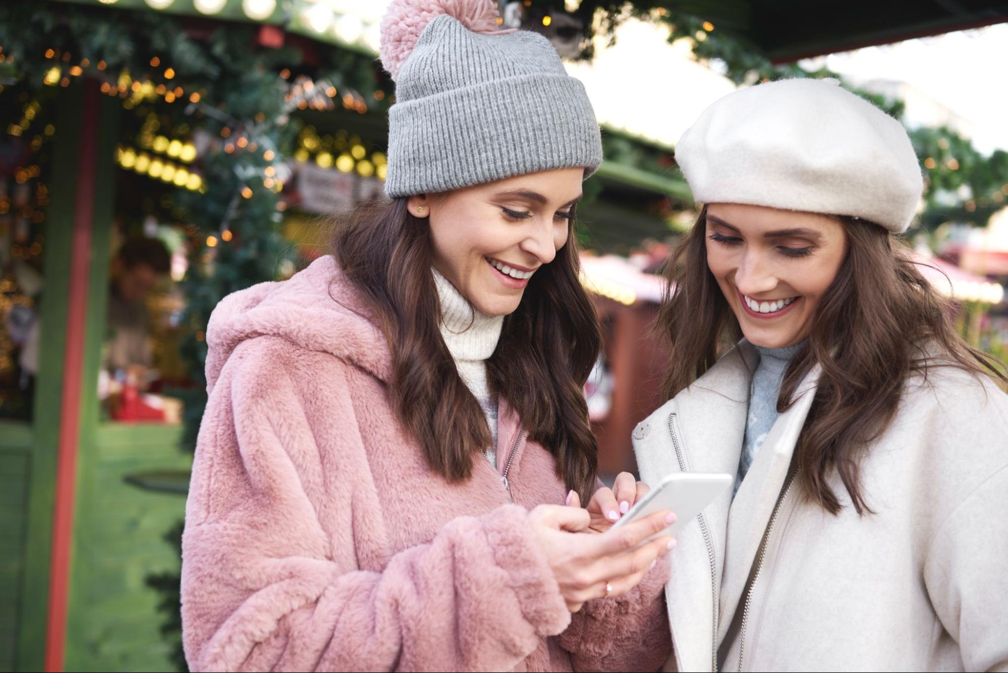 Two young women in winter hats smiling at a smartphone outdoors