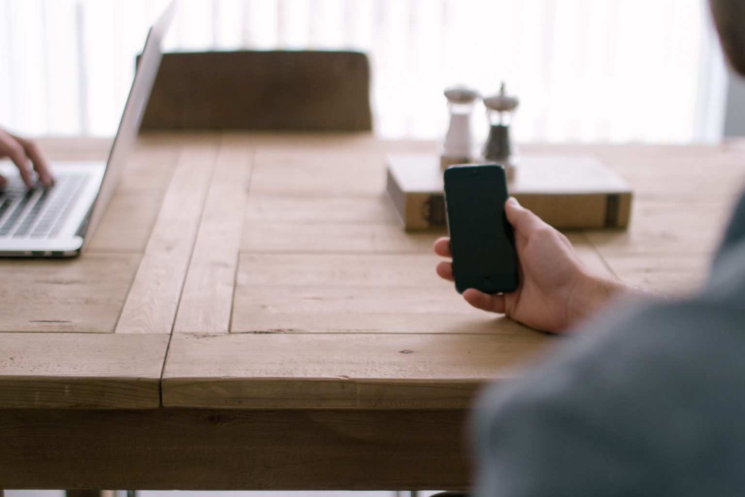 Person holding a smartphone at a wooden table beside a laptop