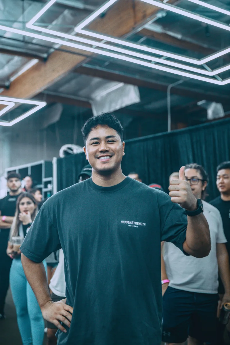 Smiling man in a black T-shirt with Hiddenstrength logo gives thumbs up indoors with people behind him.