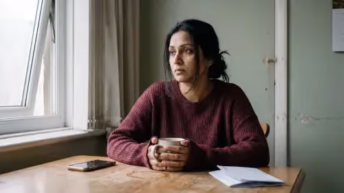 Donna seduta a un tavolo di cucina con una tazza tra le mani e lo sguardo perso nel vuoto, in una luce mattutina grigia