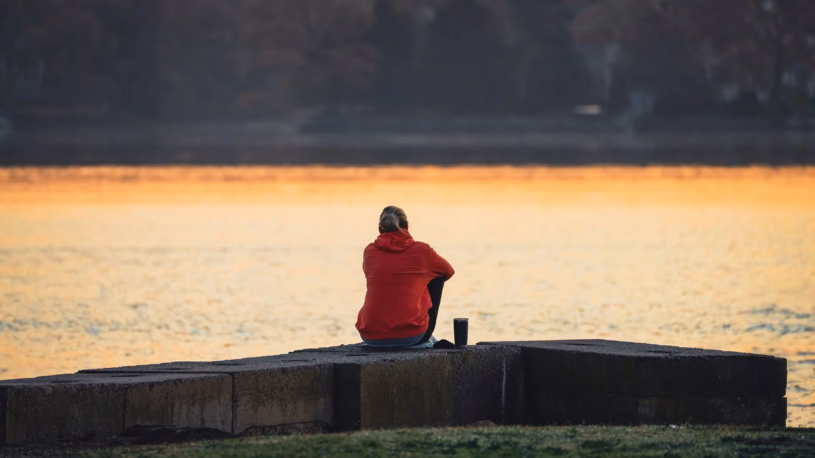 Una persona con una felpa rossa con cappuccio è seduta in riva a un lago al tramonto, immersa nei suoi pensieri in serena solitudine. — Cara Denison – Pexels