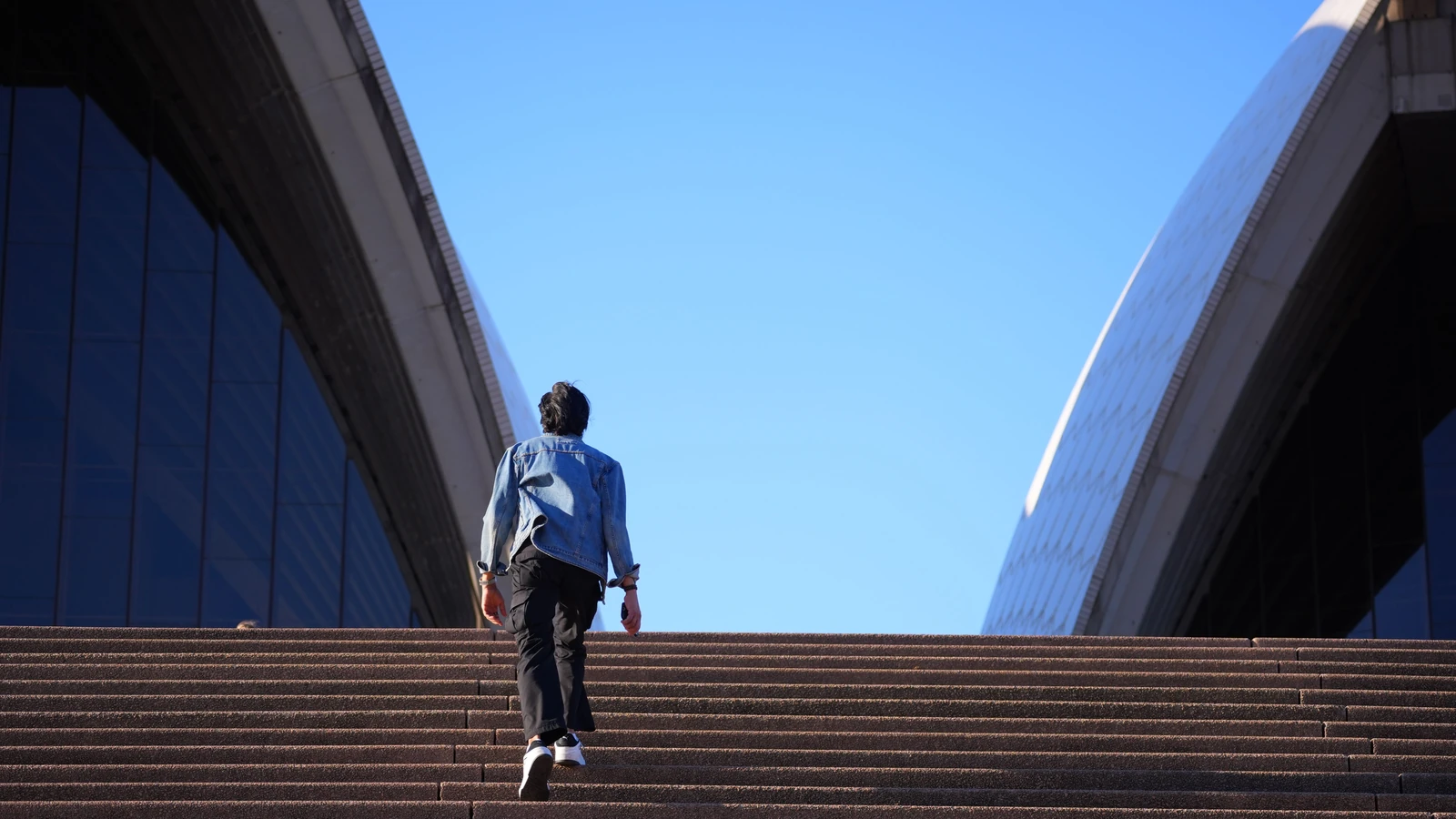 Persona che cammina verso l'Opera House di Sydney sotto un cielo terso e azzurro.