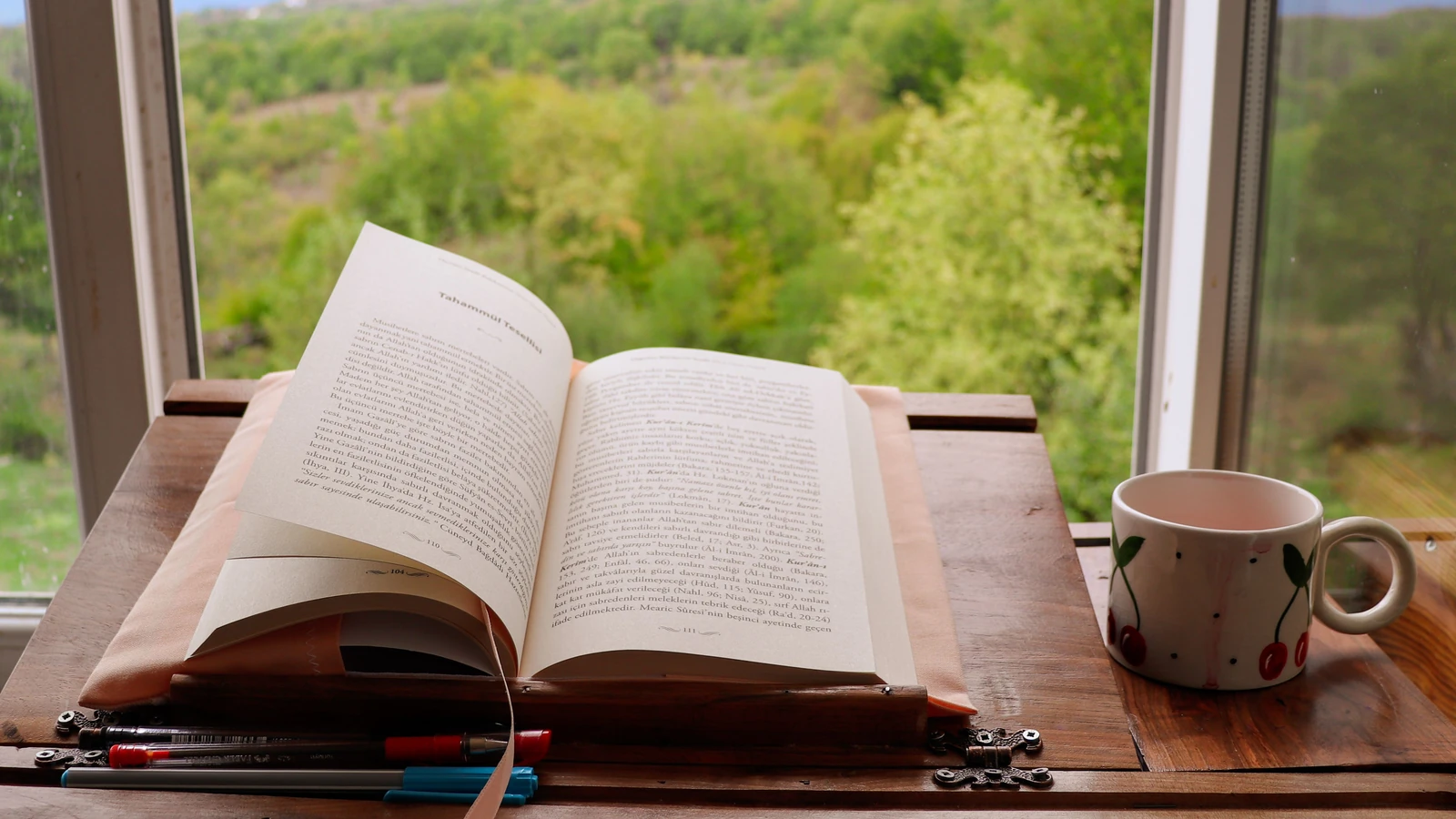 Un libro aperto e una tazza da caffè con un motivo a ciliegie su un tavolo vicino a una finestra panoramica.