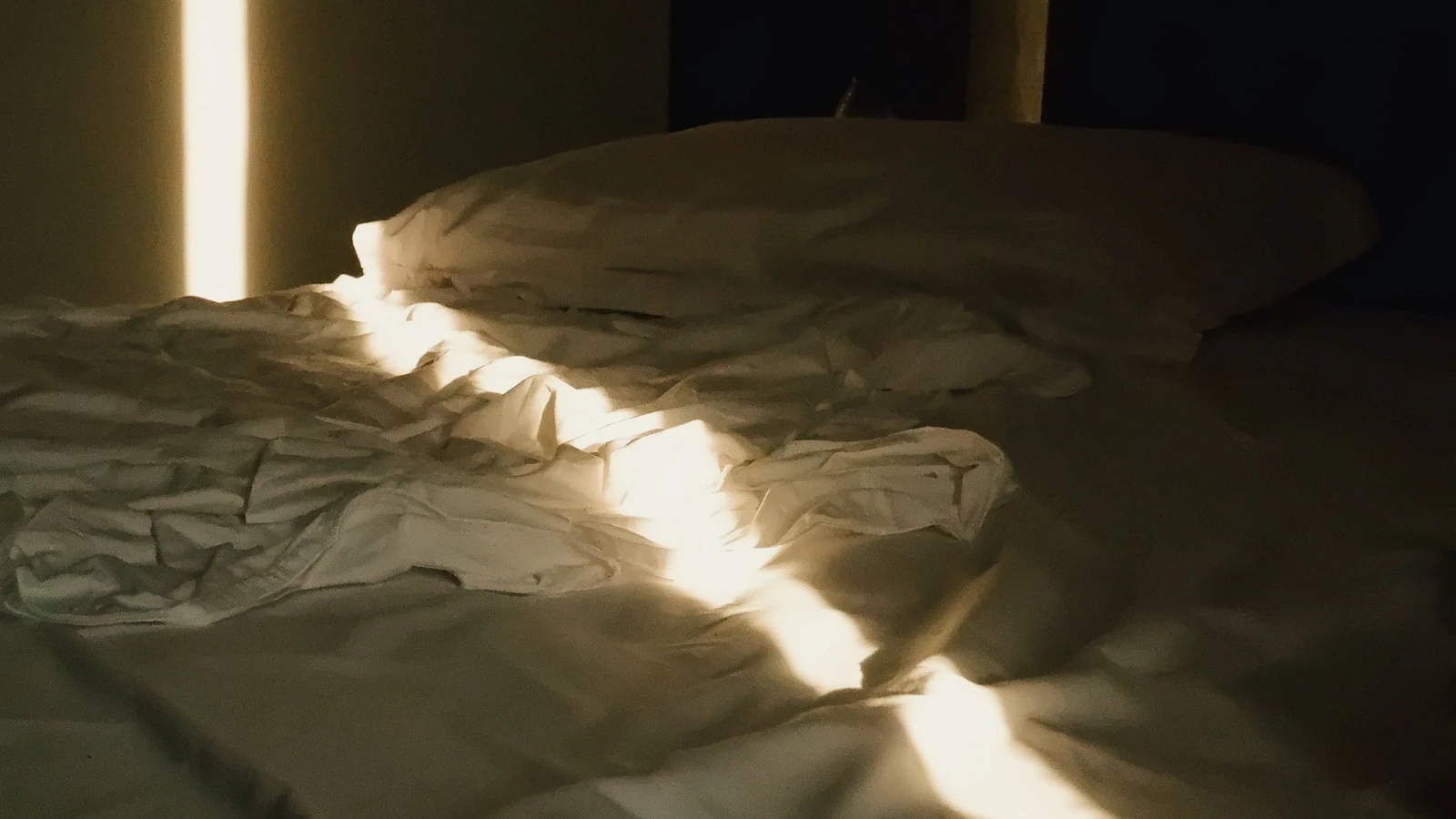 La tenue luce del sole che filtra dalla finestra illumina un letto sfatto, creando una tranquilla atmosfera mattutina.