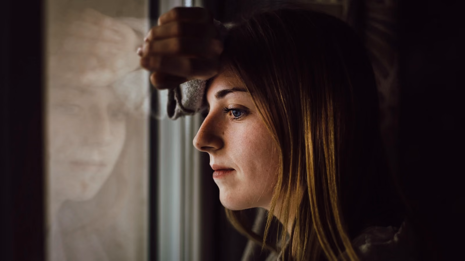 Una giovane donna contemplativa guarda fuori da una finestra con un'espressione riflessiva, all'interno. — Juan Pablo Serrano – Pexels