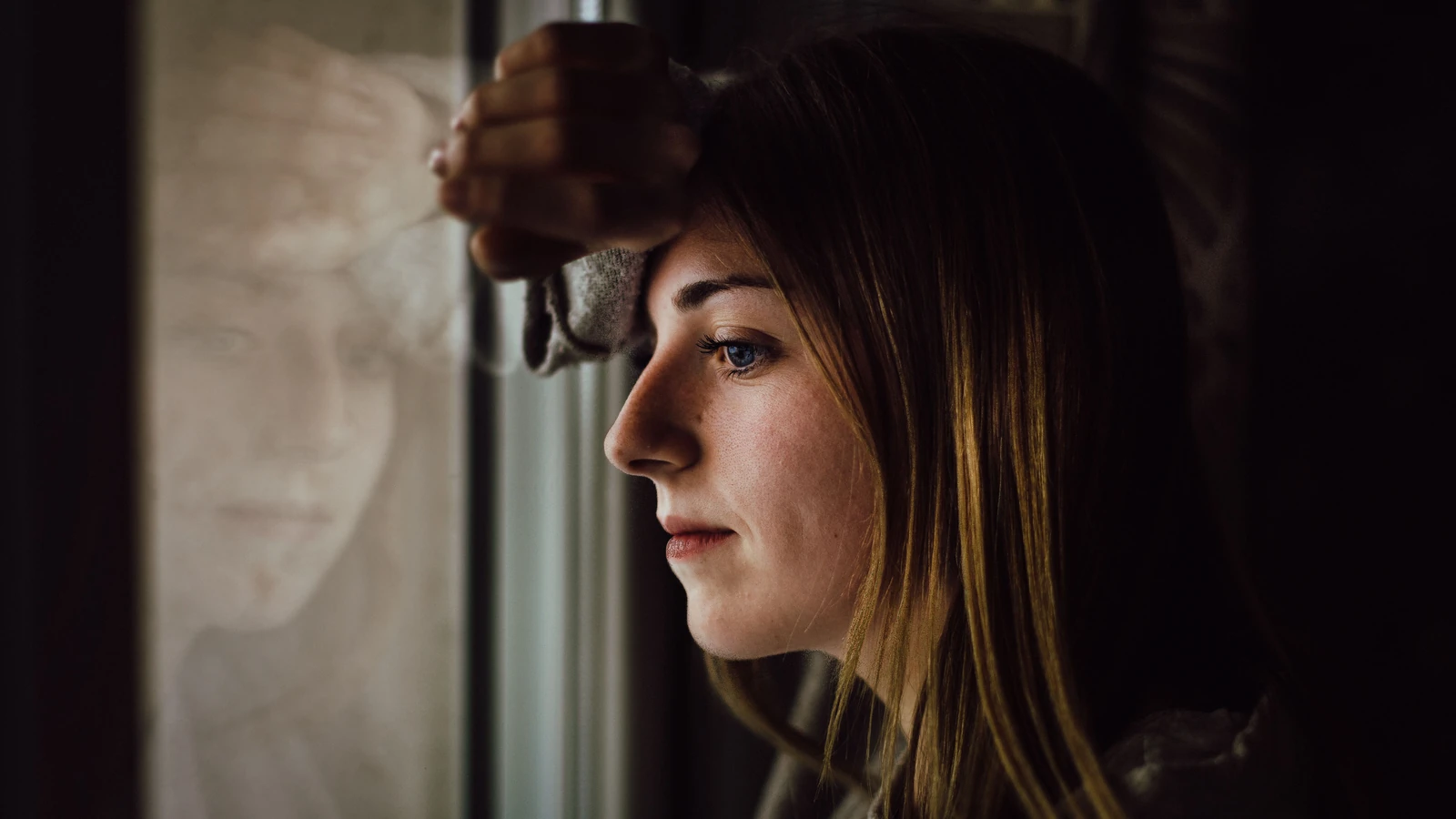 Una giovane donna contemplativa guarda fuori da una finestra con un'espressione riflessiva, all'interno. — Juan Pablo Serrano – Pexels